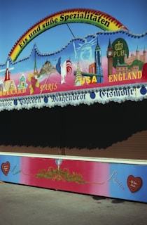 A colorful stall with a sign advertising ice cream and sweets in German. The top is decorated with illustrations of landmarks from various countries like the Eiffel Tower and Big Ben. Below, there are captions for countries like Holland, Paris, USA, and England. The colors are vibrant, with a gradient background and small candy cane motifs.