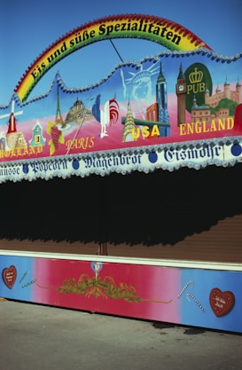 A colorful stall with a sign advertising ice cream and sweets in German. The top is decorated with illustrations of landmarks from various countries like the Eiffel Tower and Big Ben. Below, there are captions for countries like Holland, Paris, USA, and England. The colors are vibrant, with a gradient background and small candy cane motifs.