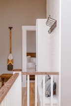 A bright and minimalistic interior space is depicted, featuring a hallway with white shiplap walls and natural wood accents. The floor is light wood, complementing the wooden railing topped with a beige, cushioned bench below. Several coat hooks are mounted on the wall, and an adjacent open doorway leads into a room with a neatly arranged bed. A decorative paddle is hung on the opposite wall.