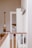 A bright and minimalistic interior space is depicted, featuring a hallway with white shiplap walls and natural wood accents. The floor is light wood, complementing the wooden railing topped with a beige, cushioned bench below. Several coat hooks are mounted on the wall, and an adjacent open doorway leads into a room with a neatly arranged bed. A decorative paddle is hung on the opposite wall.