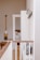 A bright and minimalistic interior space is depicted, featuring a hallway with white shiplap walls and natural wood accents. The floor is light wood, complementing the wooden railing topped with a beige, cushioned bench below. Several coat hooks are mounted on the wall, and an adjacent open doorway leads into a room with a neatly arranged bed. A decorative paddle is hung on the opposite wall.