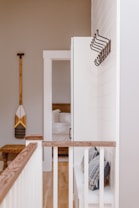 A bright and minimalistic interior space is depicted, featuring a hallway with white shiplap walls and natural wood accents. The floor is light wood, complementing the wooden railing topped with a beige, cushioned bench below. Several coat hooks are mounted on the wall, and an adjacent open doorway leads into a room with a neatly arranged bed. A decorative paddle is hung on the opposite wall.