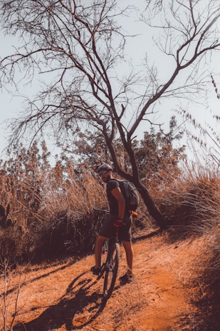 A person wearing a helmet and backpack is standing next to a mountain bike on a dirt path in a dry, arid landscape. Leafless branches of a tree stretch across the sky, with tall grass and shrubs in the background. The lighting suggests late afternoon with warm tones dominating the scene.
