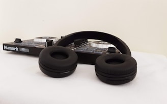 A pair of black over-ear headphones lying on a surface next to a Numark DJ controller. The controller features several buttons and dials, with a logo visible on the side. The setting appears minimalistic with a neutral background.