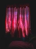 A dimly lit room features red curtains with intricate patterns, illuminated by light filtering through. Shadows enhance the texture of the fabric, creating a dramatic contrast against the dark atmosphere. A hint of a bed or seating area is visible at the bottom.
