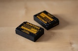 Two rechargeable camera batteries are placed on a wooden surface. The batteries are black with yellow labeling and branding details, indicating that they are Patona brand batteries designed for LP-E12 models.