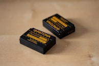 Two rechargeable camera batteries are placed on a wooden surface. The batteries are black with yellow labeling and branding details, indicating that they are Patona brand batteries designed for LP-E12 models.