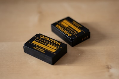 Two rechargeable camera batteries are placed on a wooden surface. The batteries are black with yellow labeling and branding details, indicating that they are Patona brand batteries designed for LP-E12 models.