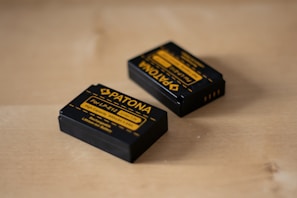 Two rechargeable camera batteries are placed on a wooden surface. The batteries are black with yellow labeling and branding details, indicating that they are Patona brand batteries designed for LP-E12 models.
