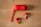 A pair of red wireless earbuds and a matching red charging case are placed on a light wooden surface. The case is oval-shaped with a wrist strap attached, lying beside the two earbuds.