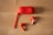 A pair of red wireless earbuds and a matching red charging case are placed on a light wooden surface. The case is oval-shaped with a wrist strap attached, lying beside the two earbuds.