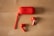 A pair of red wireless earbuds and a matching red charging case are placed on a light wooden surface. The case is oval-shaped with a wrist strap attached, lying beside the two earbuds.