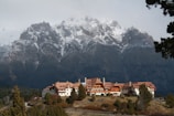 A large lodge-style building with a red-tiled roof is nestled in front of towering mountains capped with snow. The structure is surrounded by trees and set against a backdrop of rugged, dramatic peaks. The sky is overcast, adding a moody atmosphere to the scene.