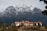 A large lodge-style building with a red-tiled roof is nestled in front of towering mountains capped with snow. The structure is surrounded by trees and set against a backdrop of rugged, dramatic peaks. The sky is overcast, adding a moody atmosphere to the scene.