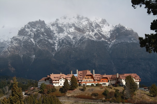 A large lodge-style building with a red-tiled roof is nestled in front of towering mountains capped with snow. The structure is surrounded by trees and set against a backdrop of rugged, dramatic peaks. The sky is overcast, adding a moody atmosphere to the scene.
