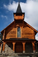 A wooden church with a pointed steeple set against a blue sky with scattered clouds. The building features log construction and a stone foundation. A stained glass window is set in the center of the upper facade.