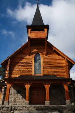 A wooden church with a pointed steeple set against a blue sky with scattered clouds. The building features log construction and a stone foundation. A stained glass window is set in the center of the upper facade.