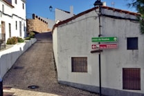 A narrow cobblestone street leads uphill between whitewashed buildings with terracotta roof tiles. A green directional sign points towards Plaza de Huelva, while a red sign indicates the direction to Iglesia Parroquial Ntra. Sra. de Consolaci&oacute;n. The walls of the buildings show signs of age with some cracks visible. A stone wall borders a part of the street, and a lone streetlamp stands against the clear blue sky.