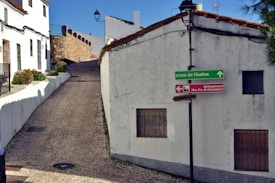 A narrow cobblestone street leads uphill between whitewashed buildings with terracotta roof tiles. A green directional sign points towards Plaza de Huelva, while a red sign indicates the direction to Iglesia Parroquial Ntra. Sra. de Consolación. The walls of the buildings show signs of age with some cracks visible. A stone wall borders a part of the street, and a lone streetlamp stands against the clear blue sky.