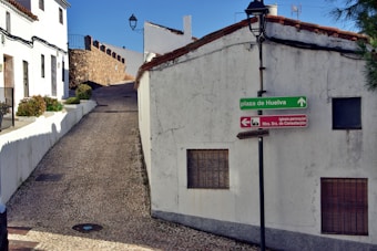 A narrow cobblestone street leads uphill between whitewashed buildings with terracotta roof tiles. A green directional sign points towards Plaza de Huelva, while a red sign indicates the direction to Iglesia Parroquial Ntra. Sra. de Consolación. The walls of the buildings show signs of age with some cracks visible. A stone wall borders a part of the street, and a lone streetlamp stands against the clear blue sky.