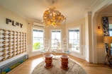 A bright and cozy room features a whimsical chandelier made of interlocking pieces and two hanging chairs by large windows. The walls are adorned with a decor element reading 'Play,' and there's a circular rug placed on hardwood floors. Two wooden stools sit in the center, enhancing the playful and inviting atmosphere.