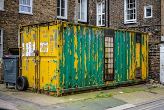 An old, weathered shipping container with peeling paint in yellow and green is situated in an urban setting. It is placed on a paved area next to a brick building with several windows. A trash bin and a discarded tire are visible beside the container. The container's surface shows signs of rust and wear, and there is a glass window with wooden framing on one side.