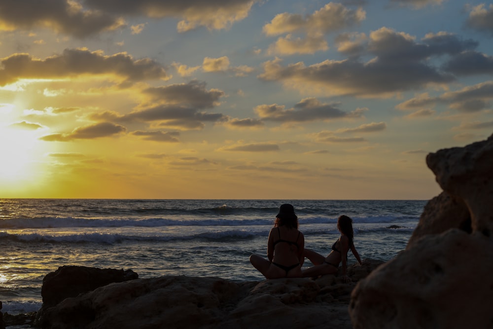 2 women sitting on rock near sea during sunset