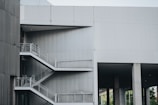 A modern architectural structure featuring an exterior metal staircase with multiple levels. The building is constructed with gray concrete and steel, and the stairs have metal railings. The overall design is minimalist and industrial, with clean lines and sharp angles.