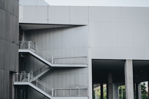 A modern architectural structure featuring an exterior metal staircase with multiple levels. The building is constructed with gray concrete and steel, and the stairs have metal railings. The overall design is minimalist and industrial, with clean lines and sharp angles.