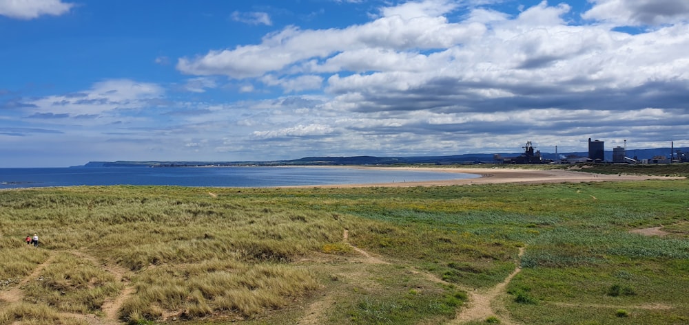 green grass field near body of water under white clouds and blue sky during daytime