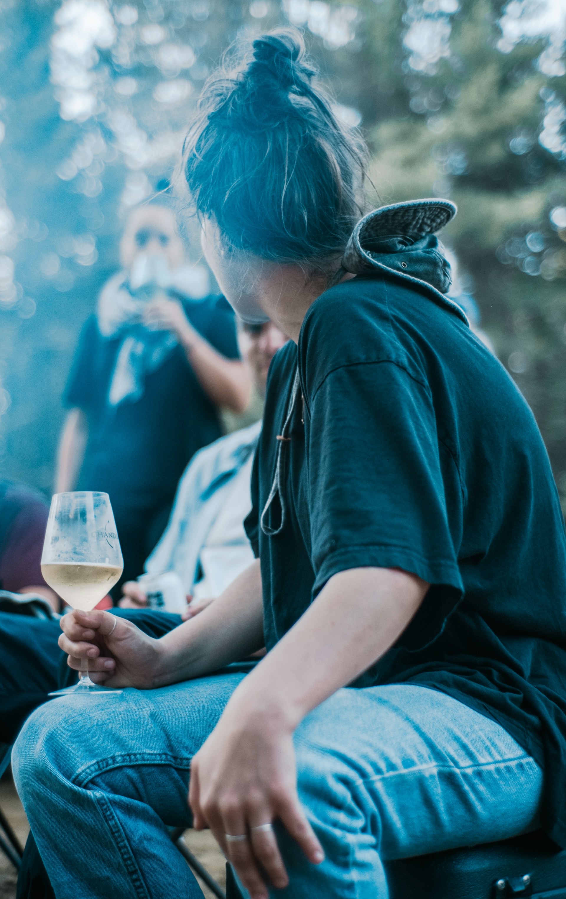 Several people are gathered outdoors, with one person prominently holding a wine glass. The setting appears relaxed and natural, with trees in the background and a soft focus on the people.