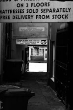 A narrow alleyway bordered by buildings with several signs advertising a store with three floors and mattress sales. Trash bins are on the left, and gates or railings are on the right, leading to a brightly lit street where a building with large windows can be seen in the background.