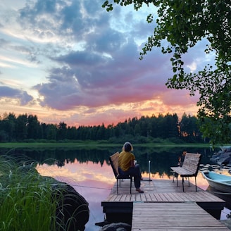man in black jacket sitting on brown wooden bench near lake during daytime