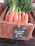 A wicker basket contains a bunch of fresh carrots with green leaves, arranged neatly. A black chalkboard sign attached to the basket displays the text 'Bunched Carrot £1.25 each', with 'Spain' written above it, indicating the origin.