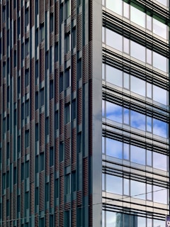 Modern high-rise buildings with glass windows reflecting the sky and surrounding structures. The left building has a facade with vertical lines and a pattern of windows, while the right side features a sleek glass exterior.