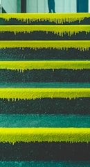 A series of steps with vibrant yellow paint dripping down the edges. The background shows part of a wall and a partial shadow or reflection of a person at the top of the stairs.