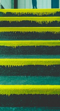 A series of steps with vibrant yellow paint dripping down the edges. The background shows part of a wall and a partial shadow or reflection of a person at the top of the stairs.