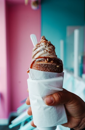 A hand holding a dessert featuring soft serve ice cream on top of a pastry cone, with caramel drizzle and nuts on top. The cone is wrapped in a white napkin, and the background displays a colorful setting with pink and teal walls.