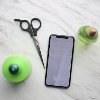 A smartphone with a reflective screen displaying a pattern is placed on a light, marble-textured surface. Nearby, there is a pair of scissors with black handles and two small green bottles, one with a blue top and the other with a beige cork.