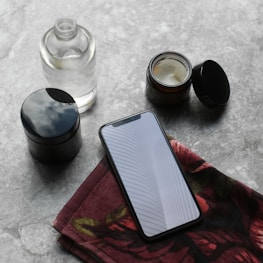 A smartphone with a reflective screen is placed on a red floral cloth next to a small open jar of cream and a clear bottle. The arrangement sits on a textured gray surface.