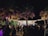 A lively outdoor nighttime gathering with a large crowd of people socializing under palm trees. String lights illuminate the area, creating a festive atmosphere. A bar or kiosk is visible in the background, adding to the party setting.