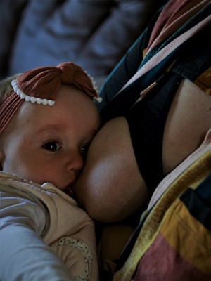 A baby wearing a headband with a bow is nursing. The baby is looking directly at the camera. The setting appears to be intimate and close, with soft lighting.
