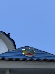 A rooftop with a blue metal siding features a circular emblem containing four colored sections in blue, yellow, green, and red. A bird is perched at the edge of a nearby white structure. The sky in the background is clear and blue.
