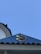 A rooftop with a blue metal siding features a circular emblem containing four colored sections in blue, yellow, green, and red. A bird is perched at the edge of a nearby white structure. The sky in the background is clear and blue.