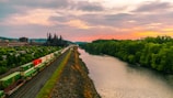 A scenic landscape features a river bordered by lush green trees on one side and a railway track on the other. The railway is carrying colorful freight containers, moving towards a large industrial facility with smokestacks in the distance. The sky is a stunning mix of orange and pink hues, indicating sunset or sunrise.