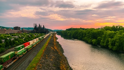 A scenic landscape features a river bordered by lush green trees on one side and a railway track on the other. The railway is carrying colorful freight containers, moving towards a large industrial facility with smokestacks in the distance. The sky is a stunning mix of orange and pink hues, indicating sunset or sunrise.