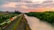 A scenic landscape features a river bordered by lush green trees on one side and a railway track on the other. The railway is carrying colorful freight containers, moving towards a large industrial facility with smokestacks in the distance. The sky is a stunning mix of orange and pink hues, indicating sunset or sunrise.