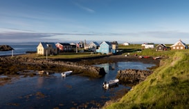 A picturesque coastal village with colorful houses resting by the seaside. There are several small boats in a small inlet surrounded by green grass and rocky shores. The sky is clear with a hint of clouds, suggesting a calm and serene atmosphere.
