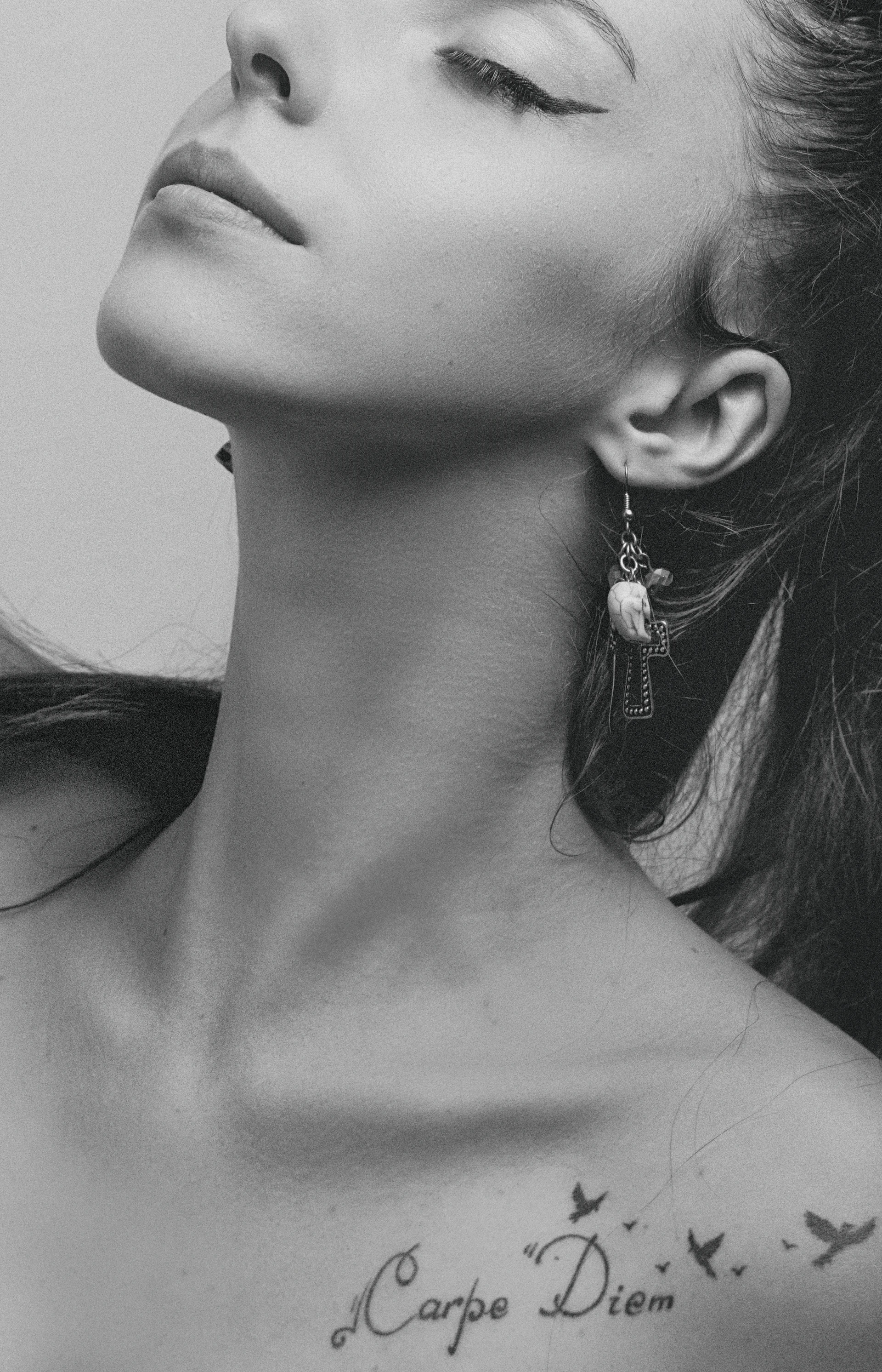 A black and white portrait of a person with a focus on the neck and lower face. The individual has raised their chin slightly, displaying an elegant posture. An earring with a unique design is visible, and there is a tattoo that says 'Carpe Diem' with small bird silhouettes near the collarbone.
