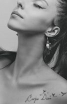 A black and white portrait of a person with a focus on the neck and lower face. The individual has raised their chin slightly, displaying an elegant posture. An earring with a unique design is visible, and there is a tattoo that says 'Carpe Diem' with small bird silhouettes near the collarbone.